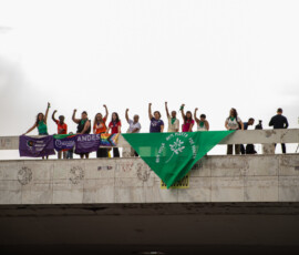 Mulheres em cima de um viaduto com bandeiras e lenços vermelhos. Ao fundo, o céu está nublado.
