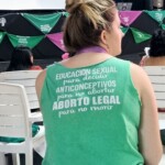 Ativista argentina na assembleia sobre Aborto, realizada no 15º EFLAC. Foto: equipe NPNM. Mulher loira com cabelos aparece de costas com uma camiseta onde diz, em espanhol: Educação sexual para decidir, anticoncepcionais para não engravidar, aborto seguro para não morrer. Ao fundo há um palco decorado com lenços verdes e rosas.