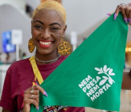 Mulher negra, de cabelos curtos e loiros, com camiseta cor bordô e brincos grandes amarelos e pretos, segura o lenço da campanha nem presa nem morta.