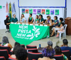 Foto de um auditório, com uma mesa no fundo, onde estão sentadas 10 pessoas. Há um lenço verde grande sobre a mesa onde se lê Nem Presa Nem Morta