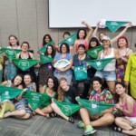 O 15º EFLAC foi uma oportunidade de intercâmbio sobre Justiça Reprodutiva, entre ativistas de diversos países da América Latina e Caribe Foto: equipe NPNM. Várias mulheres, algumas em pé e outras sentadas no chão, seguram lenços verdes e sorriem. Elas estão em uma sala com paredes beges e há uma tela de projeção ao fundo.