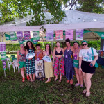 Grupo de brasileiras no espaço de Oficinas do 15º EFLAC. Foto: equipe NPNM. Oito mulheres posam, em pé, em frente a uma estrutura de barraca branca, que está decorada com lambes e lenços da campanha NPNM.