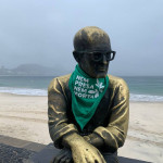A estátua de Carlos Drummond de Andrade, em Copacabana, recebeu um adereço da luta pela legalização do aborto, no amanhecer do dia 28 de Setembro. Foto: Amanda Gabriel. Foto da estátua de Carlos Drummond de Andrade vestindo um lenço verde. Ao fundo, a praia de Copacabana.