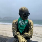 A estátua de Carlos Drummond de Andrade, em Copacabana, recebeu um adereço da luta pela legalização do aborto, no amanhecer do dia 28 de Setembro. Foto: Amanda Gabriel. Foto da estátua de Carlos Drummond de Andrade vestindo um lenço verde. Ao fundo, a praia de Copacabana.