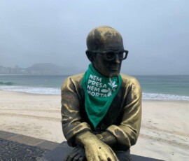 A estátua de Carlos Drummond de Andrade, em Copacabana, recebeu um adereço da luta pela legalização do aborto, no amanhecer do dia 28 de Setembro. Foto: Amanda Gabriel. Foto da estátua de Carlos Drummond de Andrade vestindo um lenço verde. Ao fundo, a praia de Copacabana.