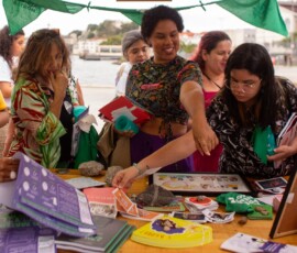 Foto de uma mesa, que fazia parte da barraca de Justiça Reprodutiva, onde á materiais diversos dispostos. Algumas mulheres observam e pegam os materiais que estão sobre a mesa.