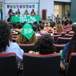 Os lenços da Campanha Nem Presa Nem Morta foram distribuídos para todes participantes, que utilizaram como adereço nos cabelos, nos braços e no pescoço. Foto: Daniele Fagundes. Imagem de um auditório. Vemos as costas de mulheres sentadas e, ao fundo, uma mesa coberta com um lenço verde da Campanha Nem Presa Nem Morta onde estão sentadas quatro mulheres palestrantes.