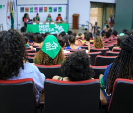 Imagem de um auditório. Vemos as costas de mulheres sentadas e, ao fundo, uma mesa coberta com um lenço verde da Campanha Nem Presa Nem Morta onde estão sentadas quatro mulheres palestrantes.