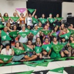 Ativistas pelo direito ao aborto marcaram presença na Assembleia sobre Justiça Reprodutiva que debateu, durante dois dias, os desafios e estratégias de luta. Foto: equipe NPNM. Várias mulheres posam em frente a um palco. Algumas estão em cima do palco, outras estão em pé no chão e outras sentadas no chão. Todas seguram lenços verdes com mensagens diversas.