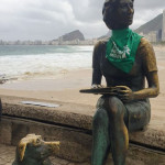 A estátua de Clarice Lispector, na praia do Leme, recebeu um adereço da luta pela legalização do aborto no amanhecer do dia 28 de Setembro. Foto: Amara Barroso. Foto da estátua de Clarice Lispector vestindo um lenço verde da campanha NPNM. Ao fundo está a praia do Leme.
