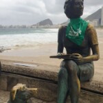 A estátua de Clarice Lispector, na praia do Leme, recebeu um adereço da luta pela legalização do aborto no amanhecer do dia 28 de Setembro. Foto: Amara Barroso. Foto da estátua de Clarice Lispector vestindo um lenço verde da campanha NPNM. Ao fundo está a praia do Leme.