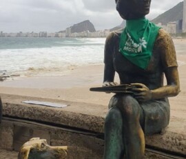 A estátua de Clarice Lispector, na praia do Leme, recebeu um adereço da luta pela legalização do aborto no amanhecer do dia 28 de Setembro. Foto: Amara Barroso. Foto da estátua de Clarice Lispector vestindo um lenço verde da campanha NPNM. Ao fundo está a praia do Leme.