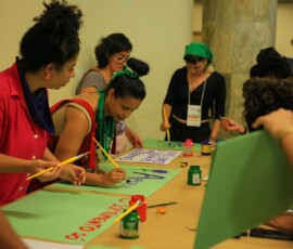 Fotos de mulheres trabalhando sobre uma mesa. Elas pintam cartazes com pinceis e tintas.