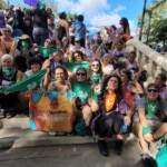 Participantes brasileiras no 15º EFLAC, reunindo-se para marchar juntas pelo Fim da Violência de Gênero, no Dia 25 de Novembro. Foto: equipe NPNM. Diversas mulheres estão sentadas em uma escada com bandeiras e lenços verdes. Ao fundo o céu azul claro e parte de um parque.