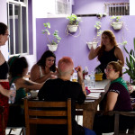No EncontrAço 2023, os trabalhos em grupo enriqueceram a elaboração de propostas para 2024 - Foto: Sandra Alves (vagaluzes.filmes) Grupo de pessoas sentadas ao redor de uma mesa com duas mulheres em pé. As pessoas estão em uma área aberta, com paredes roxas e algumas plantas.