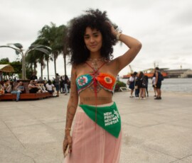 Mulher de cabeos crespos na altura do ombro, com top de crochê colorido e saia longa rosa, usa o lenço da camanha nem presa nem morta como acessório na cintura. ao fundo, imagens do Museu do Amanhã no Rio de Janeiro.