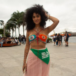 Festival WOW em close. Foto: Mari Bley. Mulher de cabeos crespos na altura do ombro, com top de crochê colorido e saia longa rosa, usa o lenço da camanha nem presa nem morta como acessório na cintura. ao fundo, imagens do Museu do Amanhã no Rio de Janeiro.