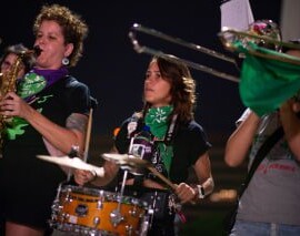 Três mulheres tocando instrumentos na batucada. Elas vestem lenços verdes da campanha Nem Presa Nem Morta.