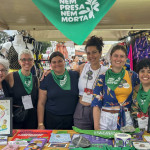 Grupo parceiro em plantão na barraca aborteira. Foto: Amara Barroso. Seis mulheres aparecem atrás de uma mesa onde estão visíveis materiais impressos diversos, como cartilhas e adesivos.