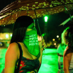 Ativista veste o lenço da Campanha Nem Presa Nem Morta durante a manifestação do dia 28 de setembro, dia latinoamericano de luta pela descriminalização do aborto. Foto: Daniele Fagundes. Foto de uma mulher de perfil,ela usa o lenço da campanha nem presa nem morta sobre o rosto, abaixo dos olhos.