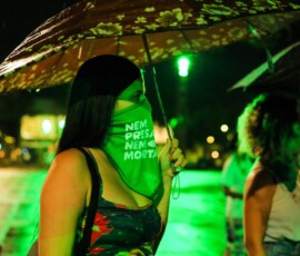 Foto de uma mulher de perfil,ela usa o lenço da campanha nem presa nem morta sobre o rosto, abaixo dos olhos.