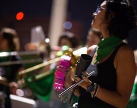 Mulheres tocando instrumentos na batucada. Elas vestem lenços verdes da campanha Nem Presa Nem Morta.