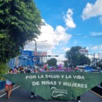 Um grupo anfitrião, da Colectiva Feminista de El Salvador, abriu ala da Marcha pelo Fim da Violência de Gênero com um lenço verde gigante. Foto: equipe NPNM. Foto de uma rua sendo ocupada por uma manifestação. Mulheres seguram um lenço verde gigante onde se lê, em espanhol: Pela saúde e pela vida de meninas e mulheres. Assina Colectiva Feminista.