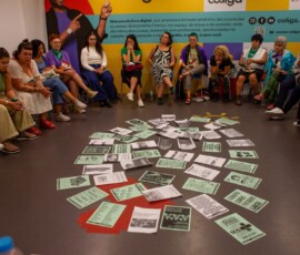 Diversas pessoas aparecem sentadas em uma roda, no centro, há diversos papéis impressos.