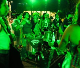 Foto de várias mulheres com instrumentos de batucada sob luz esverdeada em espaço público.