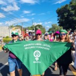 Ativistas bolivianas somando-se à Marcha pelo Fim da Violência de Gênero, com palavras de ordem pelo direito ao aborto. Foto: equipe NPNM. Foto de uma rua sendo ocupada por uma manifestação. Mulheres seguram um lenço verde grande onde se lê, em espanhol: Aborto legal, seguro e gratuito. Há um símbolo do feminismo em branco e a palavra Bolívia.