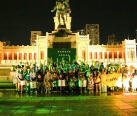Diversas pessoas com lenços verdes e outros adereços da luta pela legalização do aborto posam para foto em frente a um monumento em praça pública.