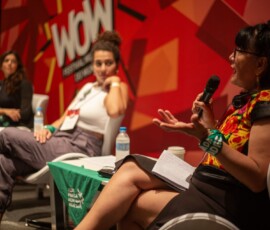 Em primeiro plano, uma mulher com microfone, atrás duas mulheres aparecem. Ao fundo, está um painel com a identidade visual do Festival WOW em vermelho.