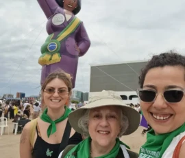 Foto de três ativistas da Nem Presa Nem Morta, com lenços do Comitê por Justiça Reprodutiva da Marcha das Mulheres Negras. Atrás, uma boneca inflável grande de pele negra, roupa roxa, faixa presidencial e punho erguido.