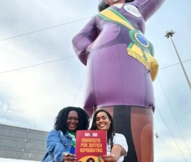 Foto com dias mulheres mostrando pra câmera a capa de um documento, que diz: Manifesto por Justiça Reprodutiva. Atrás, uma grande boneca inflável de roupa roxa e a faixa de presidenta da república.