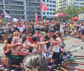 Foto de uma performance no ato Mulheres Vivas. No centro, mulheres de joelhos estão abraçadas em círculo. Elas estão meladas de tinta vermelha e vestem pernas de pau. Nas margens, uma multidão com bandeiras e cartazes assiste.