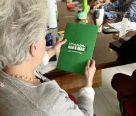 Foto de uma mulher segurando uma pasta verde da campanha Criança Não é Mãe. Ao fundo uma mesa de reunião com outras mulheres.