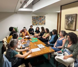 Foto de uma sala com diversas mulheres em volta de uma mesa grande em que há pastas verdes, papéis, copos e um computador. Atrás delas dois quadros na parede.