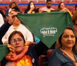 Foto com duas mulheres sentadas em um auditório, uma delas mostra o lenço da campanha Criança Não é Mãe. Atrás delas, estão outras pessoas sem foco.