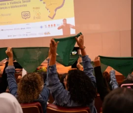 Foto pegando as costas e cabelos de mulheres sentadas em um auditório que erguem lenços da campanha Criança Não é Mãe. Ao fundo, um telão grande com a projeção da logo do evento.