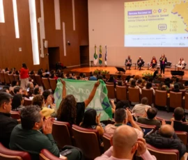 Foto ampla do auditório com pessoas sentadas e um palco grande com 5 palestrantes. Uma delas está em pé com o microfone na mão e as demais sentadas em poltronas. Atrás, um a projeção no telão da identidade visual do evento.