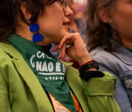 Foto em close de duas mulheres jovens. Uma delas usa óculos de grau, brincos grandes azuis, lenço verde da campanha Criança Não é Mãe no pescoço e blazer verde.