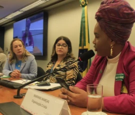 Foto de uma mulher negra de turbante e blazer rosa com o adesivo da campanha Criança Não é Mãe. Ela segura um celular e fala em um microfone de mesa, enquanto olha para o lado oposto da câmera. Na sua frente, um copo de água e uma placa pequena, que diz: Mariane Marçal - Organização Criola. Ao seu lado, três mulheres sentada à mesa. Atrás delas, uma bandeira do brasil e um telão.