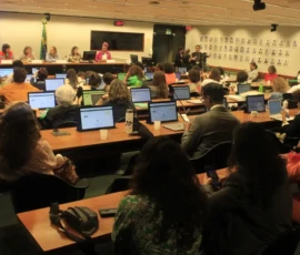 Foto tirada a partir do fundo da sala que mostra diversas pessoas sentadas em bancadas da sala de Comissões da Câmara Federal. As bancadas possuem pequenas telas e microfones pretos. Ao fundo, uma mesa com as palestrantes. Atrás delas, um telão e a banceira do Brasil. Na parede ao lado fotografia com rostos de pessoas.