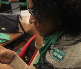 Foto do close de lado de uma mulher de cabelos pretos cacheados, óculos de grau, com um lenço verde no pescoço e um adesivo da campanha Criança Não é Mãe. Ela segura e olha fixamente para o celular. Atrás, em uma bancada, uma bolsa preta, um lenço verde e outro celular.