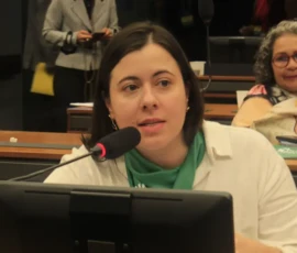 Foto da deputada Sâmia Bonfim de cabelos pretos lisos, lenço verde no pescoço e camisa branca. Ela fala em um microfone preto. À sua frente aparece parte de um monitor. Atrás dela, uma bancada com pastas verdes e uma mulher sentada.