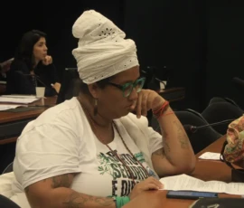 Foto de close e perfil de uma mulher negra de turbante branco, camiseta branca, colar que lê uma carta que está sobre a bancada. À frente uma bolsa de estampa colorida.