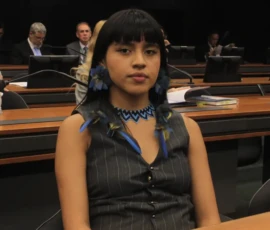 Foto de uma mulher indígenas de cabelos pretos longos, brincos de flores e penachos azuis, colar azul, blusa preta está sentada numa bancada. Atrás dela, bancadas com monitores e microfones, com algumas pessoas.
