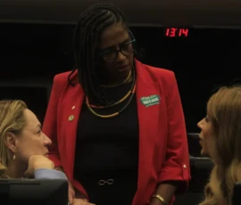 Foto da ex-parlamentar, ...., com blazer vermelho e adesivo da campanha Criança não é Mãe. Ela está em pé e conversa com a deputada Erika Hilton do PSOL/SP que está sentada. Ao lado, a deputada Delegada Adriana Accorsi do PT/GO.
