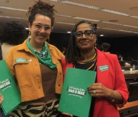 Foto com Laura Molinari, de lenço verde no pescoço, jaqueta bege, pin de arruda e adesivo no peito. Ao lado, a ex-deputada ... de blaze vermelho, adesivo e colares coloridos. Ambas seguram a pasta verde com a logo da campanha Criança Não é Mãe. Atrás delas pessoas, bancadas e monitores.