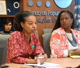 Foto em plano médio de duas representantes do Fundo de Populações das Nações Unidas, sentadas na mesa. Uma delas tem blusa com estampa vermelha e fala olhando para frente, próximo a suas mãos está um copo de água. Atrás delas, no canto direito, uma bandeira do Brasil e outra da ONU. Ao centro, a logo do Fundo na parede e uma jovem sentada no canto esquerdo.